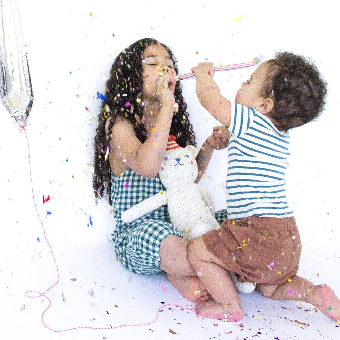 Two children playing with a stuffed animal on a white background with confetti.