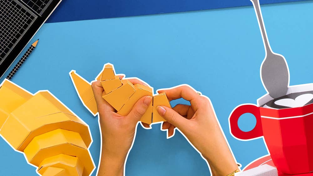 Hands assembling a yellow paper craft on a blue background with a red mug and spoon.
