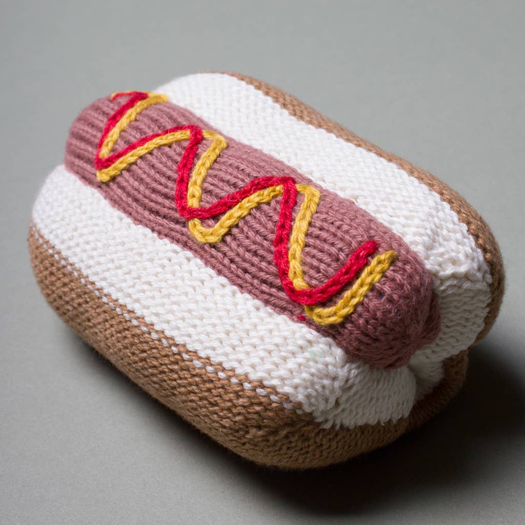 Knitted hot dog toy with brown bun, white sausage, and red and yellow mustard on a gray background