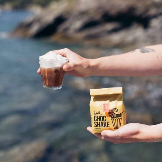 Person holding a Choc Shake bag and a glass of chocolate shake by a body of water.