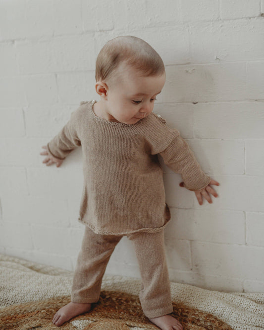 Baby in a brown outfit standing against a white brick wall.