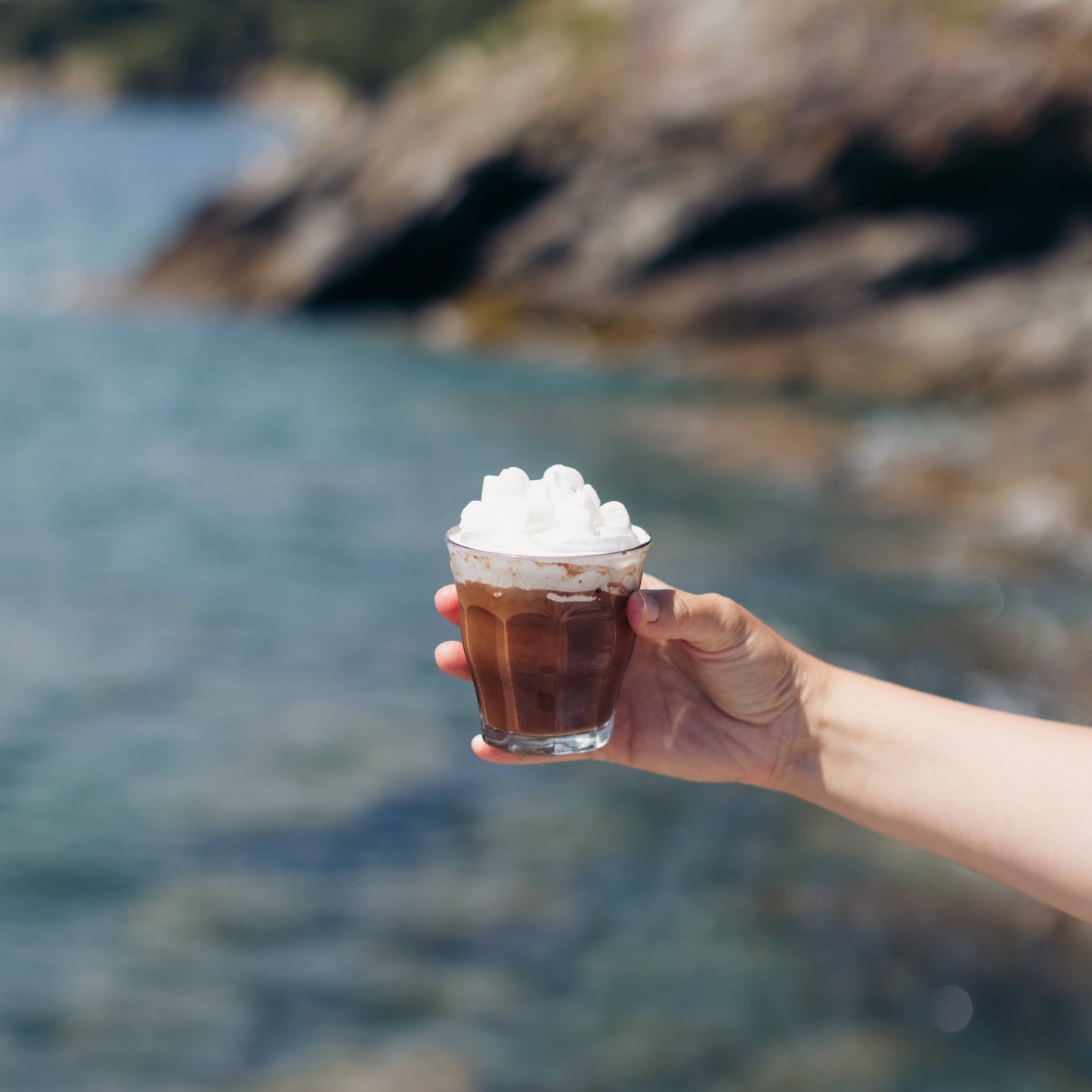 Hand holding a glass of chocolate milkshake with whipped cream against a blurred natural background