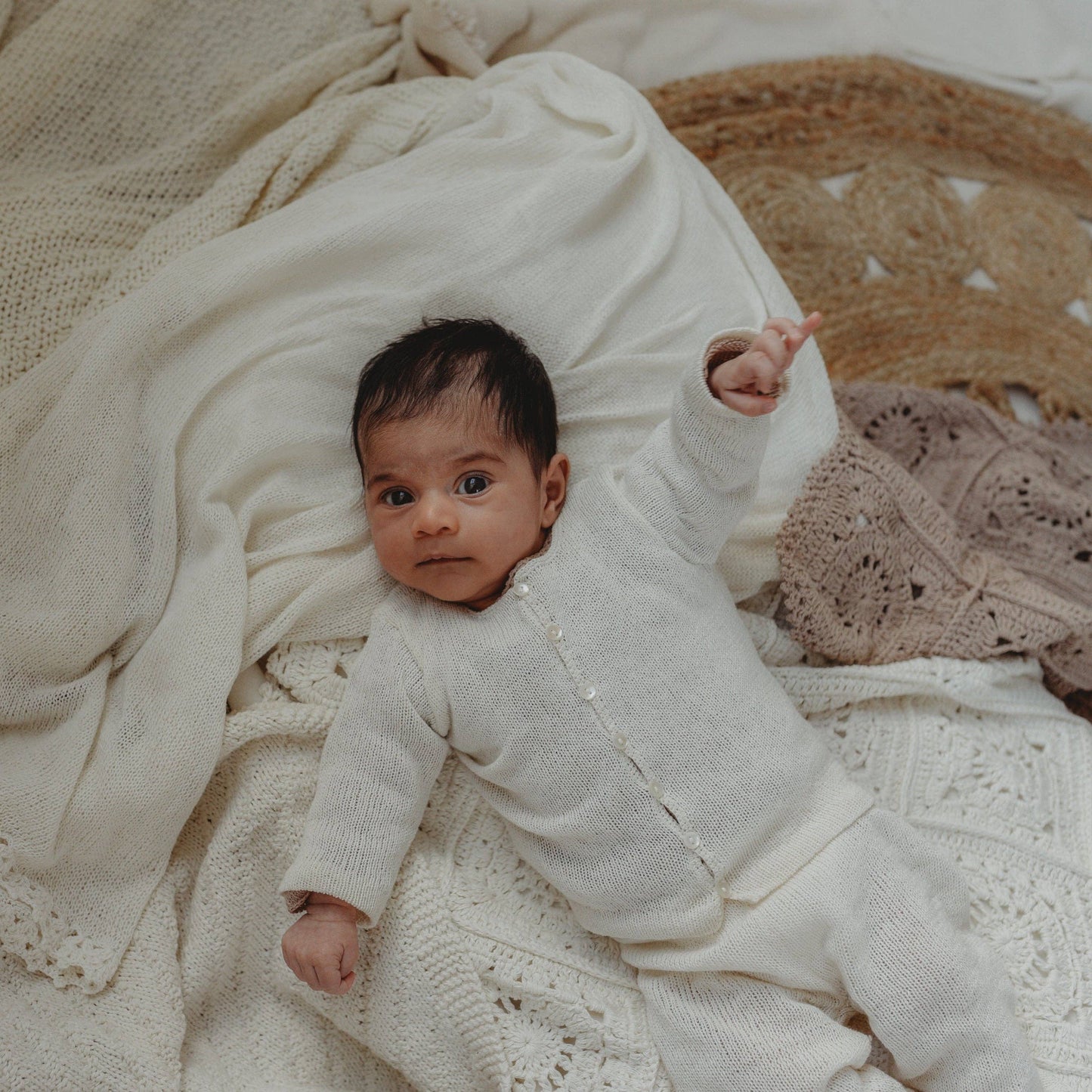 Baby in a white outfit lying on a textured blanket with a textured wall in the background