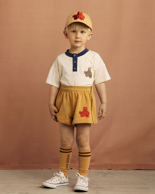 Child wearing a beige cap, white shirt with blue trim, yellow shorts, and white sneakers on a brown background