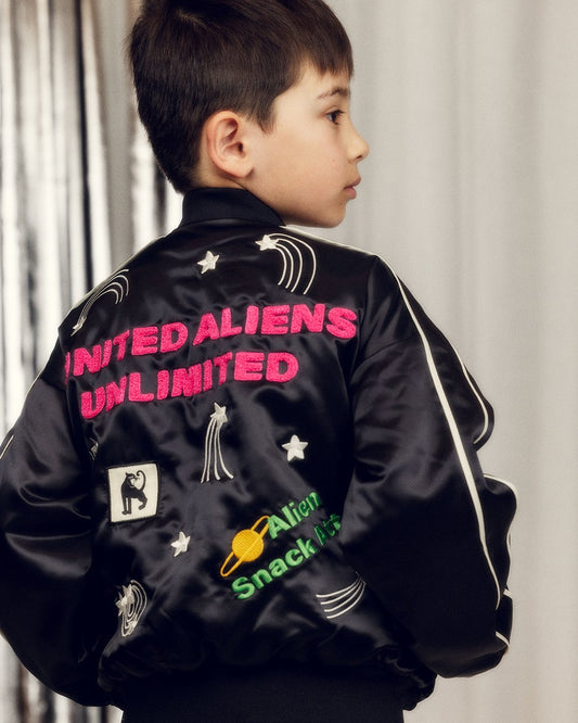 Child wearing a black jacket with colorful text and graphics, standing against a neutral background.