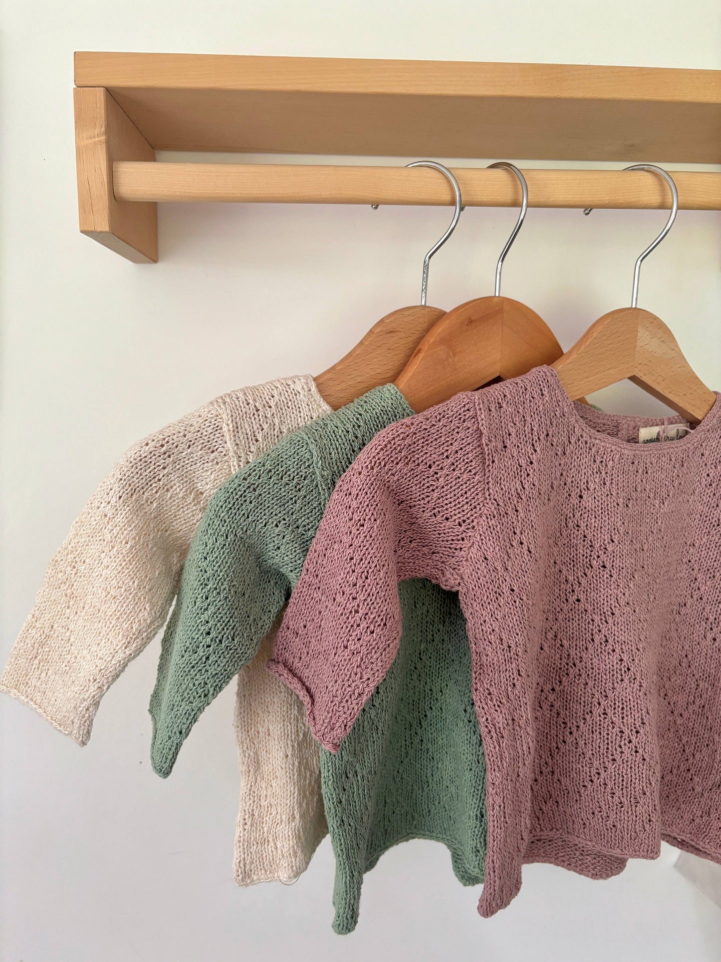 Three knitted sweaters hanging on a rack against a white wall.
