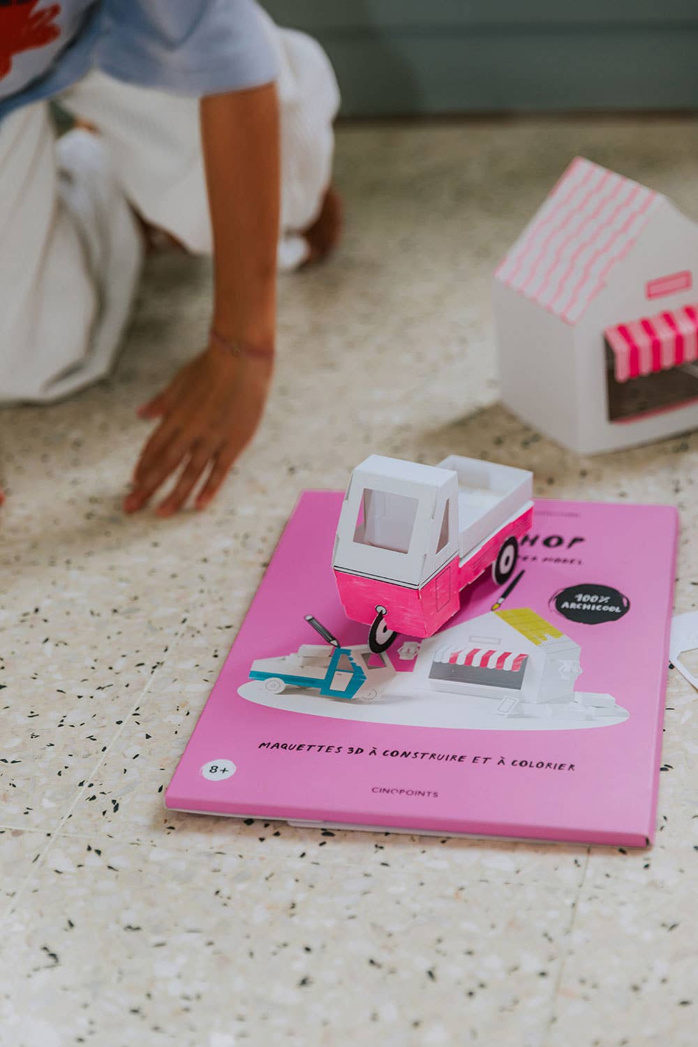 Toy ice cream truck on a pink book with a child's hand reaching towards it.