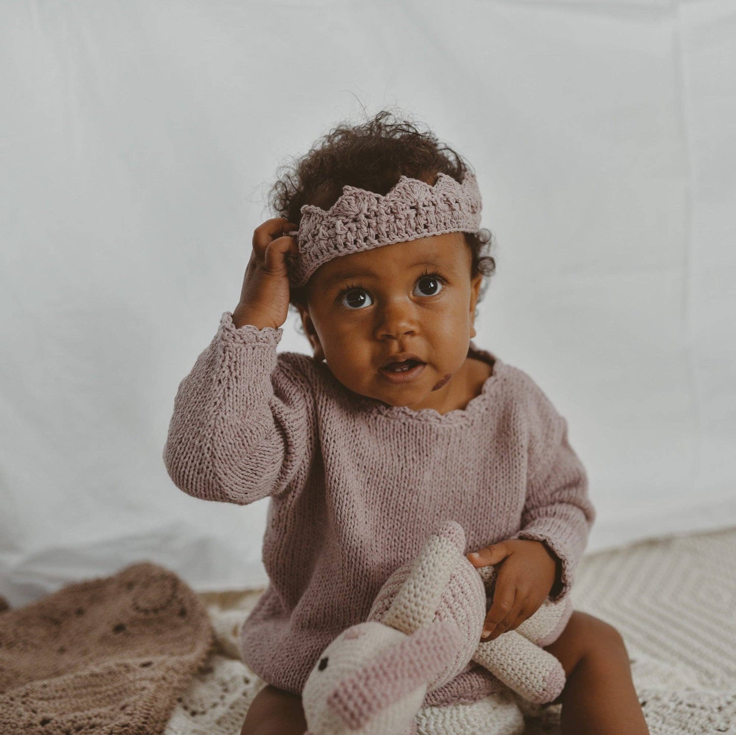 Baby wearing a pink knitted outfit and headband, holding a pink knitted toy against a white background.