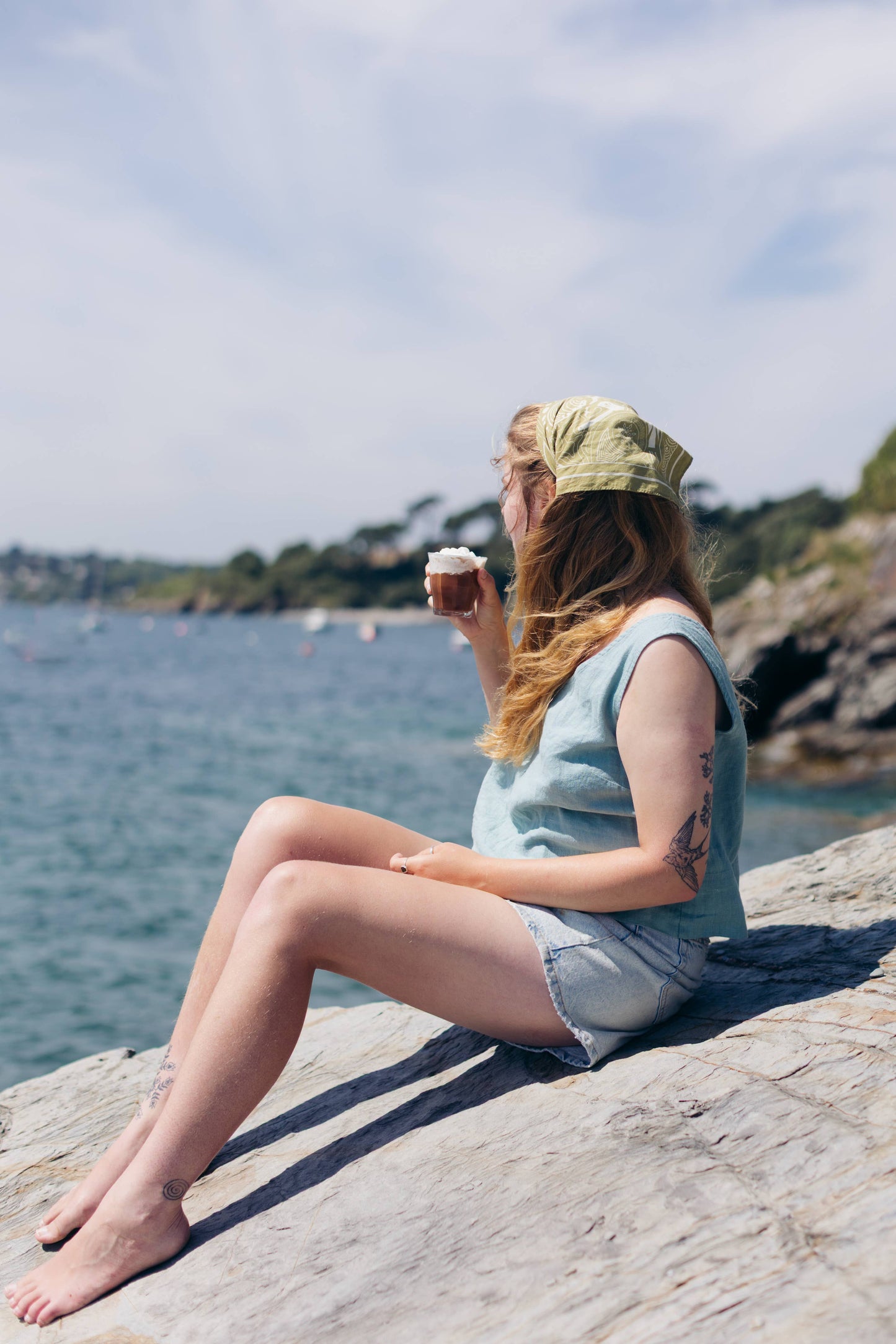 Person sitting by a body of water holding a cup, wearing a light blue sleeveless shirt and denim shorts.