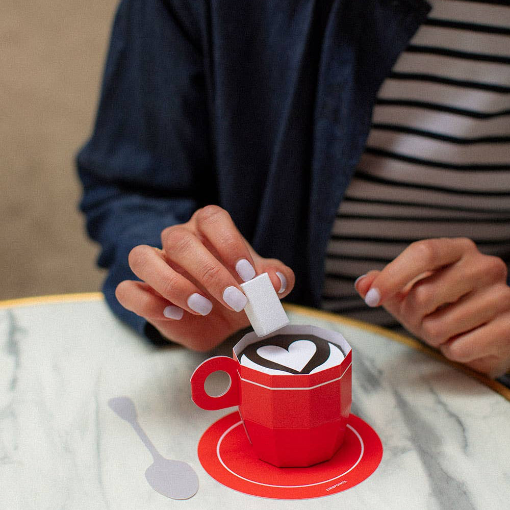Person making a heart-shaped design in coffee with a red cup on a marble table.