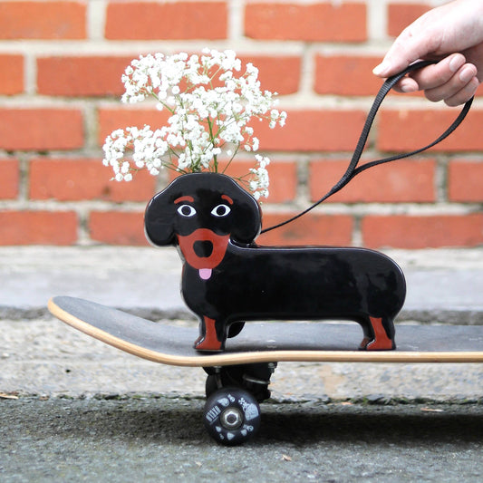 Black dachshund-shaped skateboard with a hand holding a leash against a brick wall.