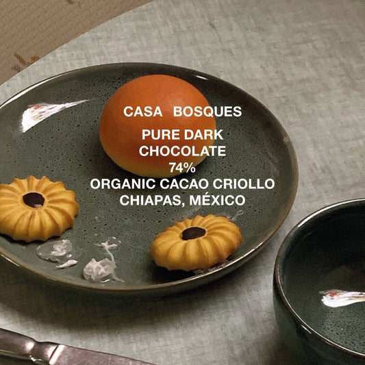 A dark chocolate bar with 74% organic cacao content from Casa Bosques, displayed on a grey plate with a whole orange and a cookie, emphasizing the flavor and quality.
