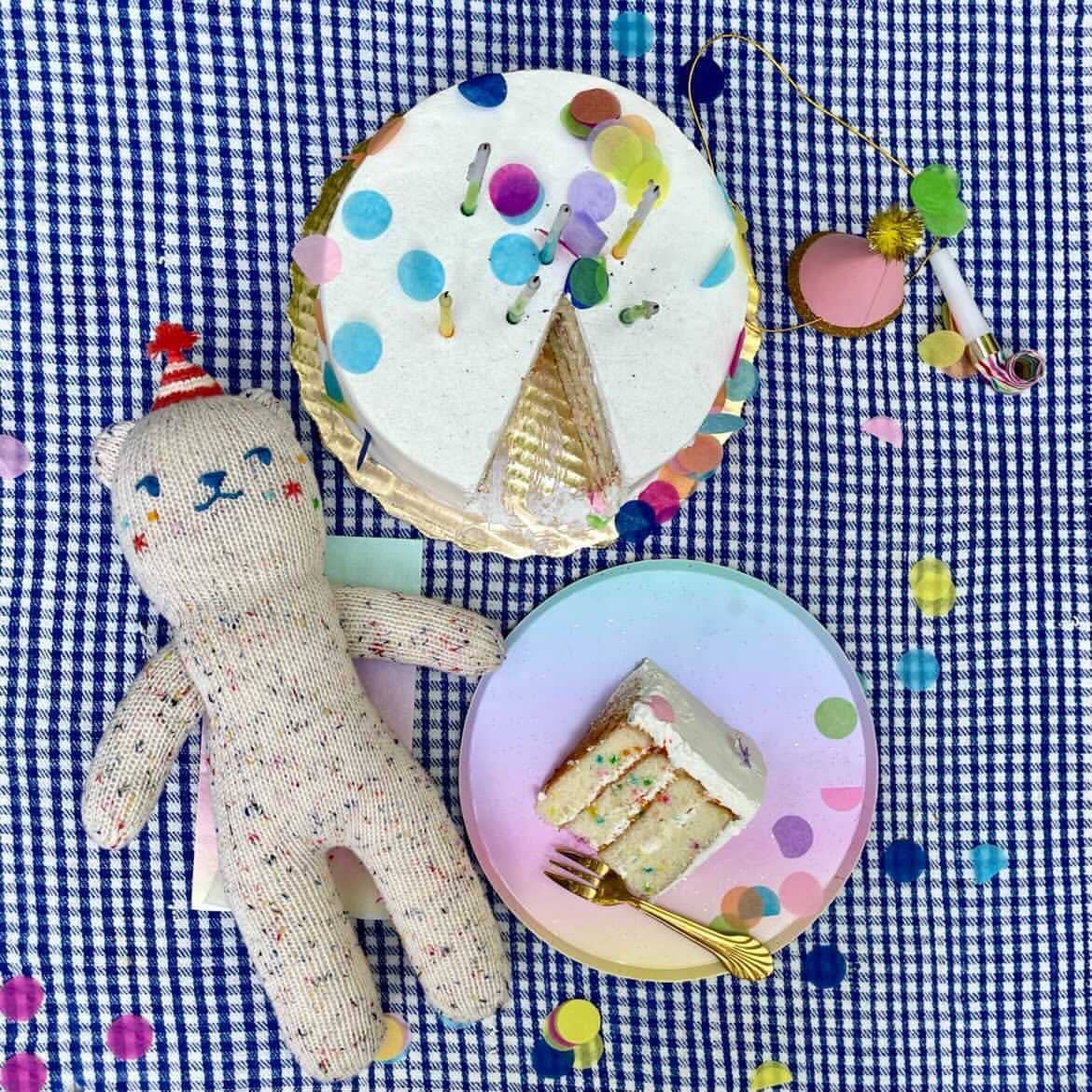 Birthday cake with candles, slice of cake on a plate, and a teddy bear on a checkered tablecloth.