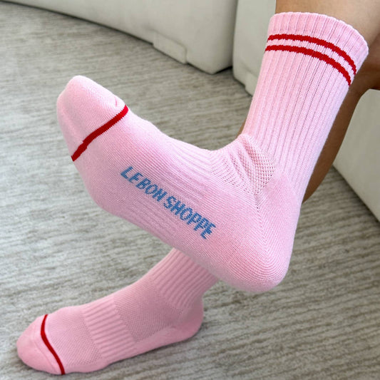A pair of Le bon shoppe amour pink boyfriend socks which are pink in colour with red stripes at the top and near the toes are worn by a person. It says Le Bon shoppe on the sole of the socks in blue colour. The floor is wooden and is beige with brown lines of detail.
