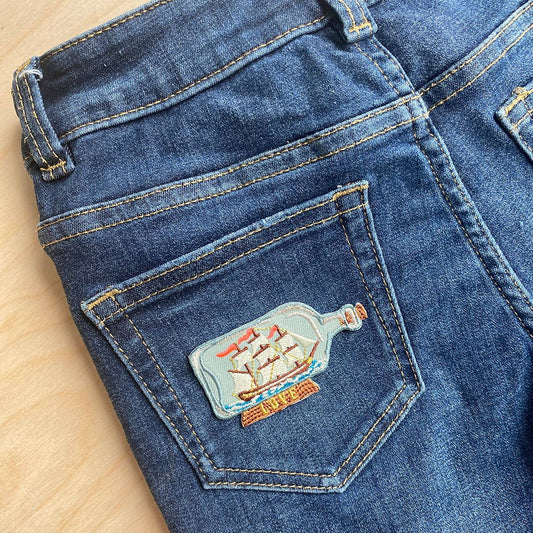 The embroidered patch applied to the back pocket of blue denim jeans, showcasing its intricate stitching, soft blue bottle, detailed ship rigging, and vintage-inspired aesthetic.