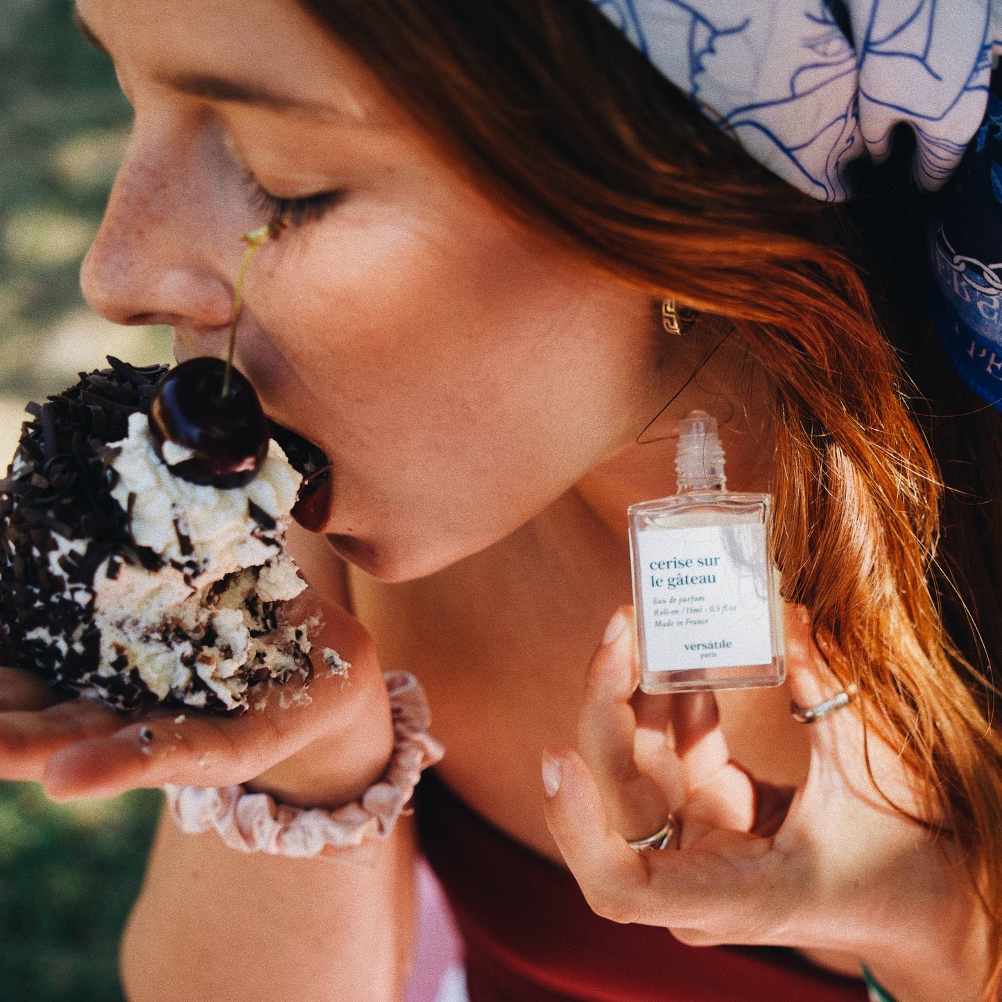 A person holding a roll-on perfume bottle labeled 'VERSATILE PARIS - CHERRY ON THE CAKE - EAU DE PARFUM' while eating a piece of cake.