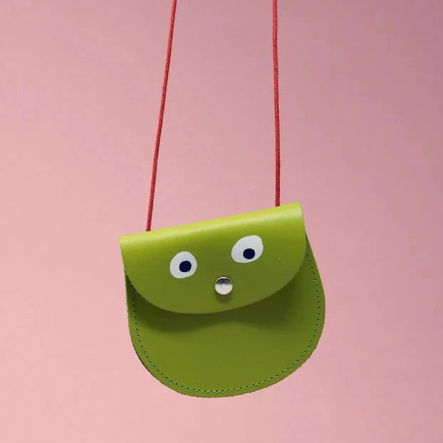 An oval shaped apple green small pocket purse with red thin rope as the bag strap hangs against a pink background. Printed onto the bag are two white googly eyes with black pupils and a small silver press button which acts as the nose and opens/closes the purse.