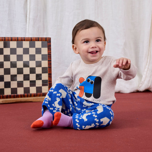 Baby sitting on a red floor wearing a white shirt with a blue dog design and blue patterned pants.