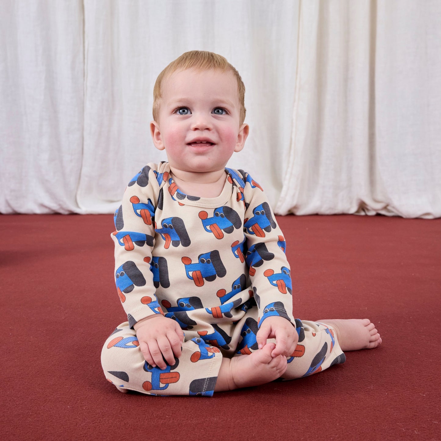 Baby sitting on a red floor wearing a patterned outfit with a white curtain background