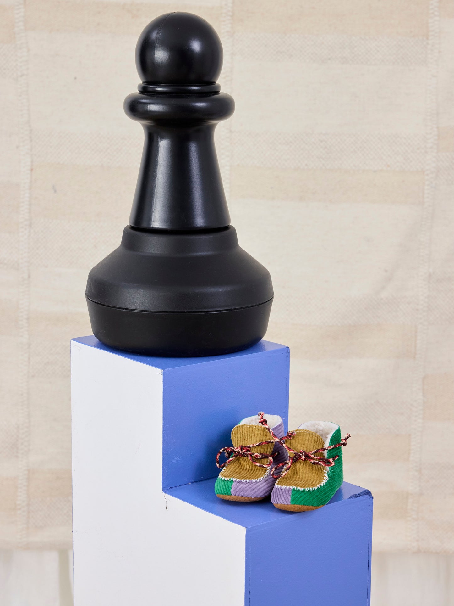 Large black chess piece on a blue platform with baby shoes below, against a beige wall.