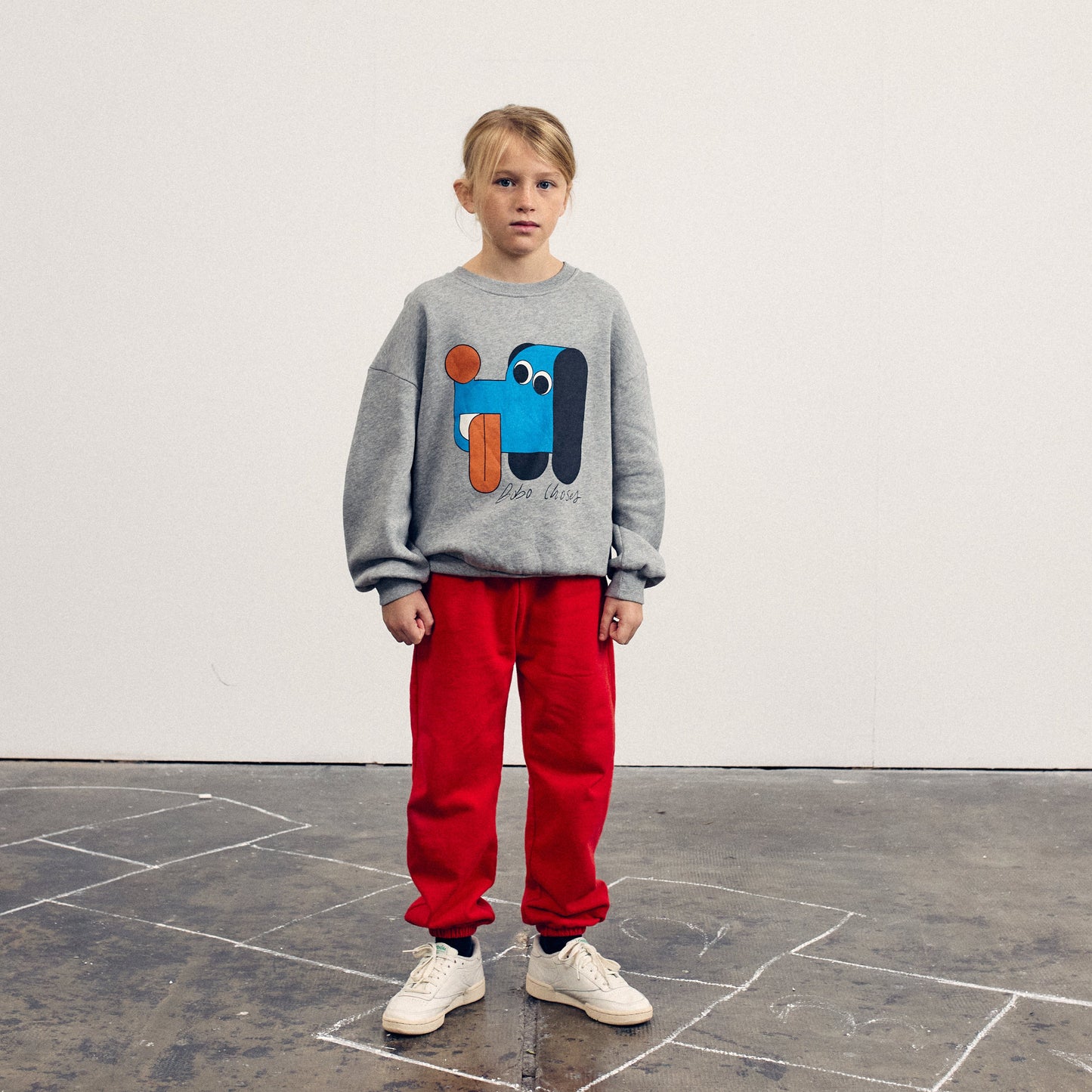 Child wearing a gray sweatshirt with a colorful dog graphic and red pants standing on a concrete floor.