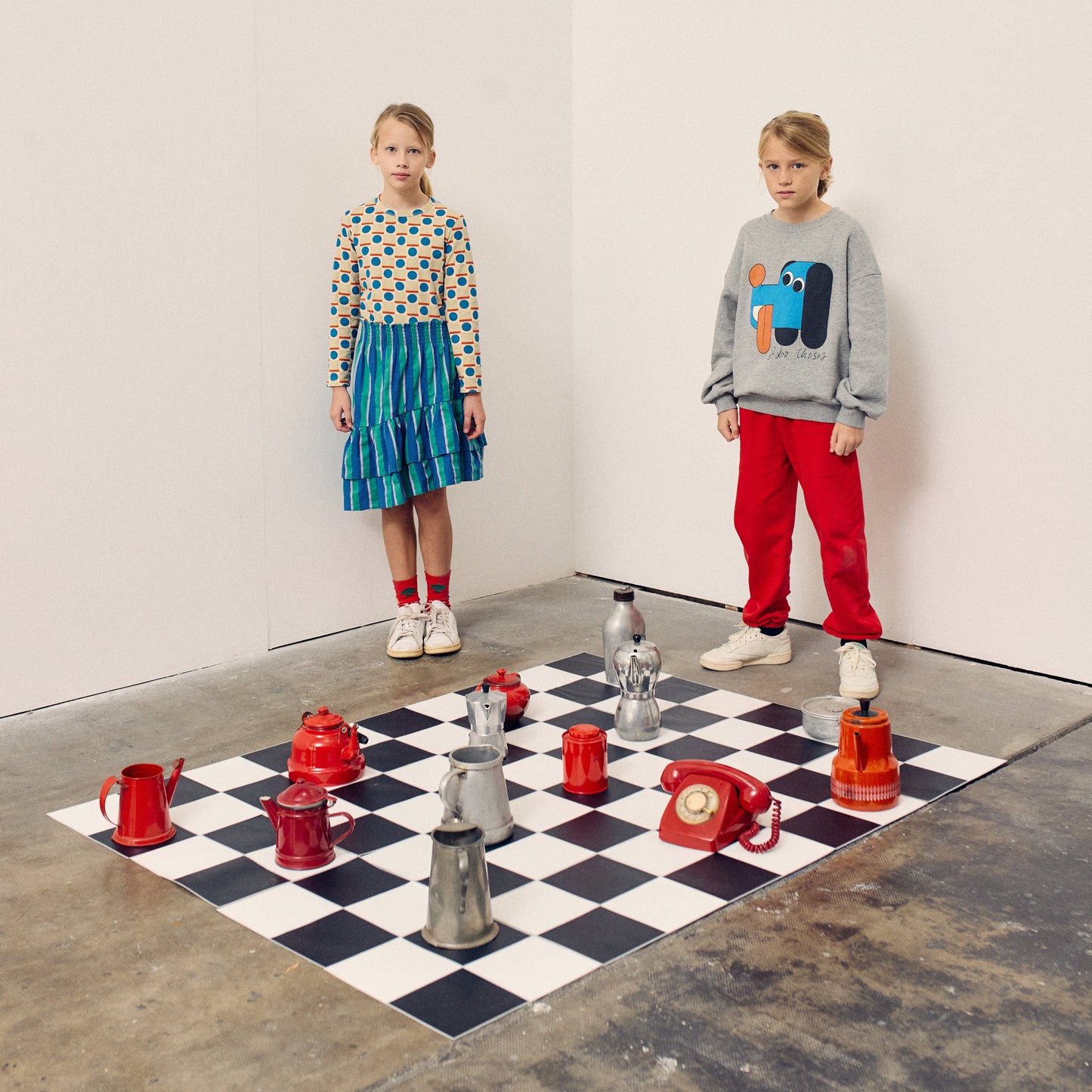 Two children standing on a checkered floor with a large chess set.