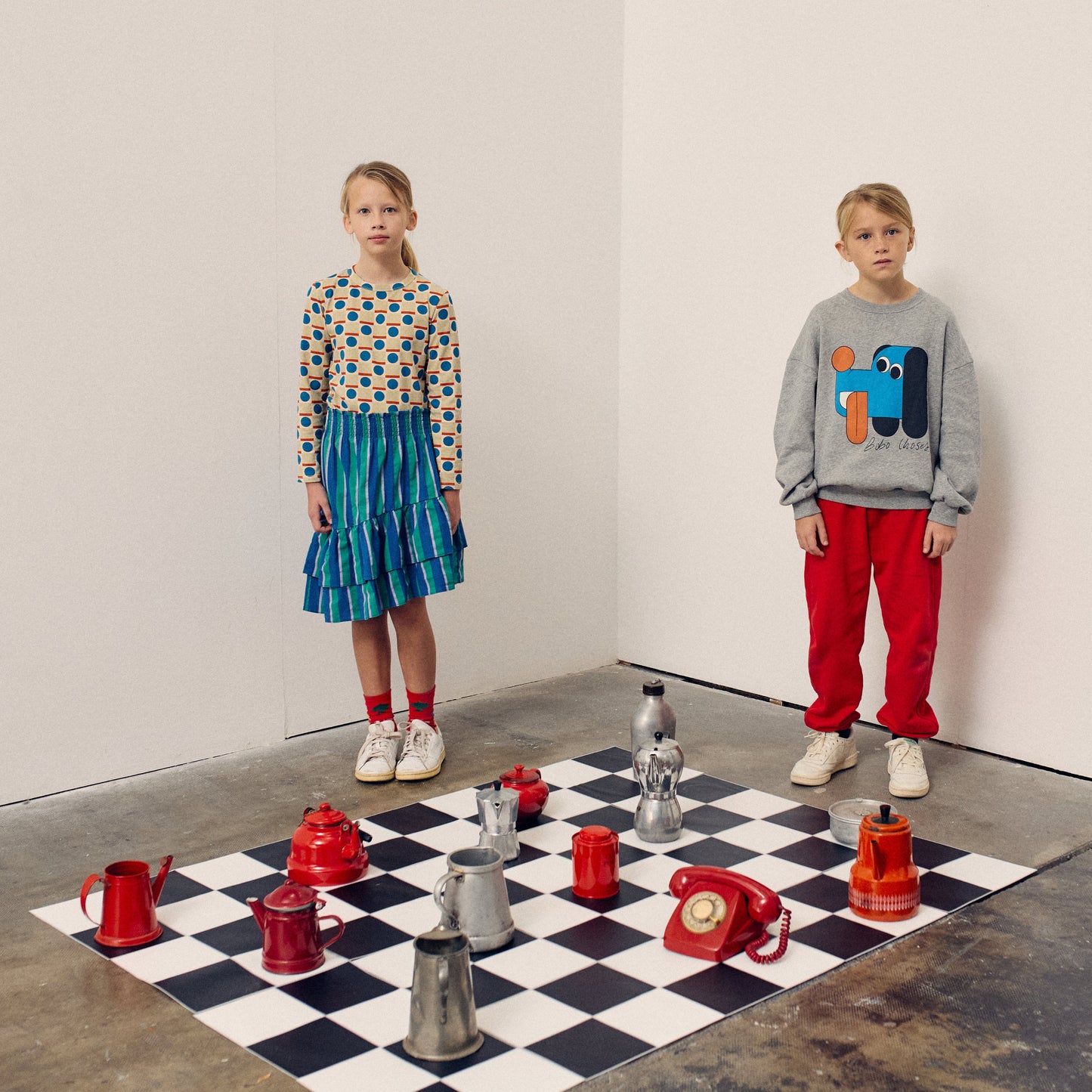 Two children standing on a checkered floor with various containers arranged in front of them.