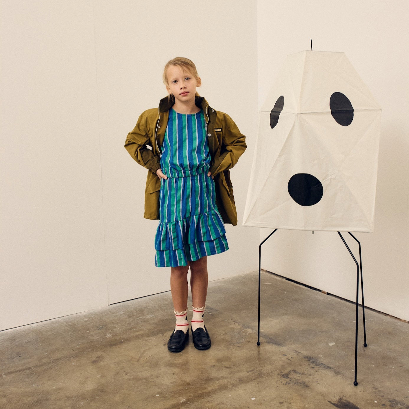 Child in a green jacket and blue dress standing next to a white wall with black dots.