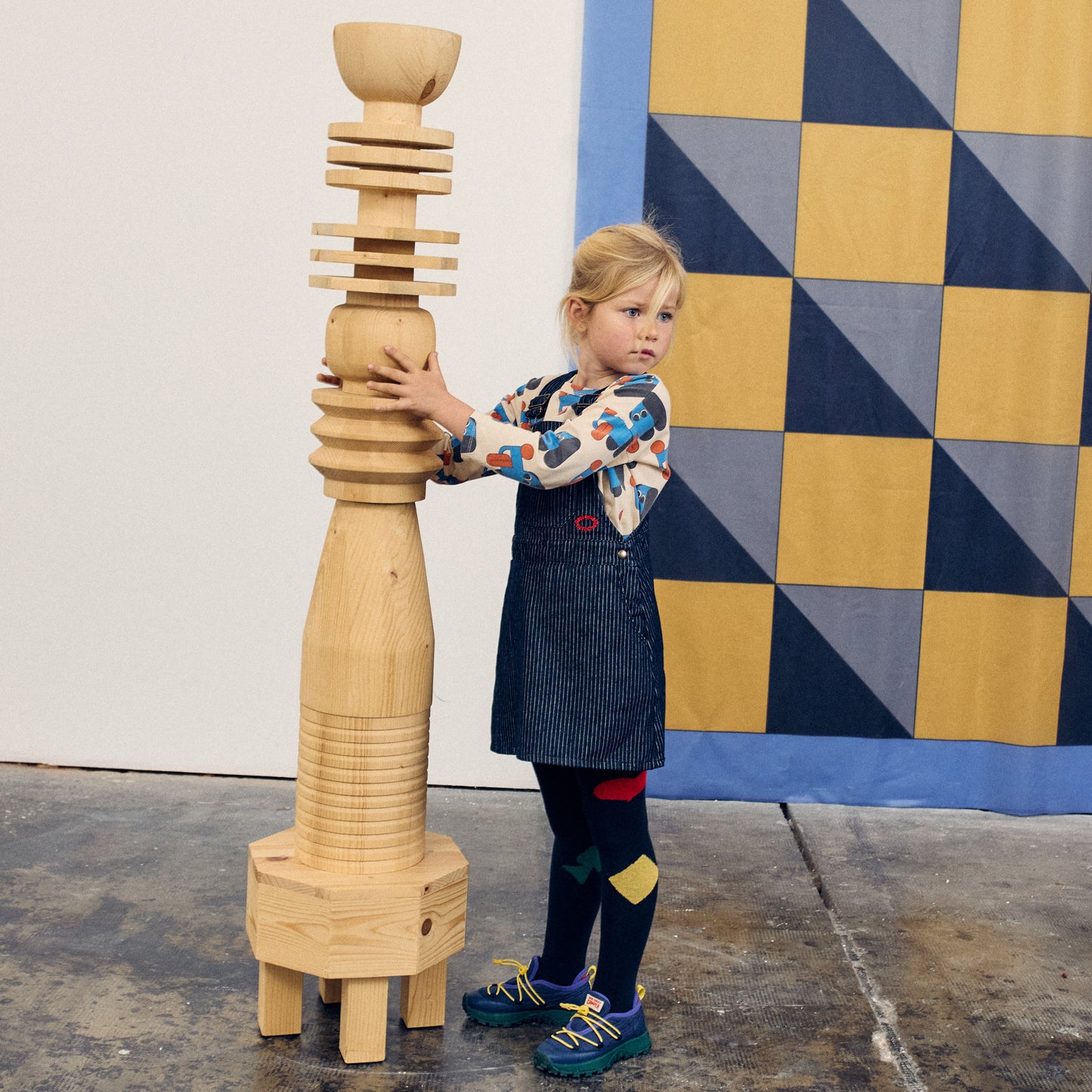 Child holding a large wooden sculpture in front of a geometric patterned wall.