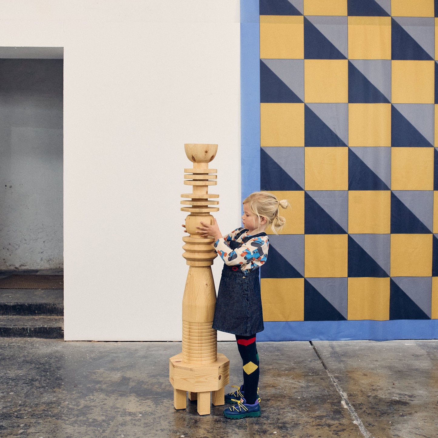 Child holding a wooden sculpture in front of a geometric patterned wall.