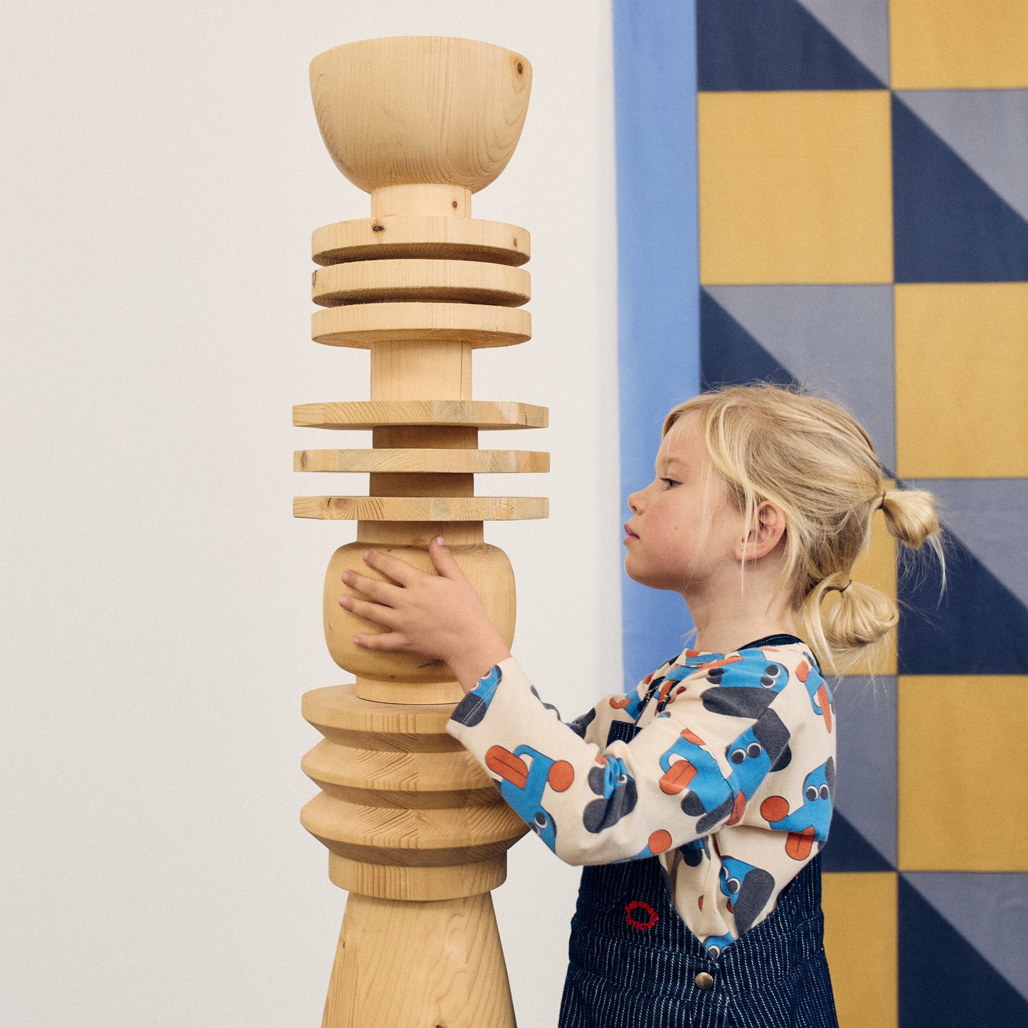 Child holding a tall wooden sculpture against a geometric-patterned wall.