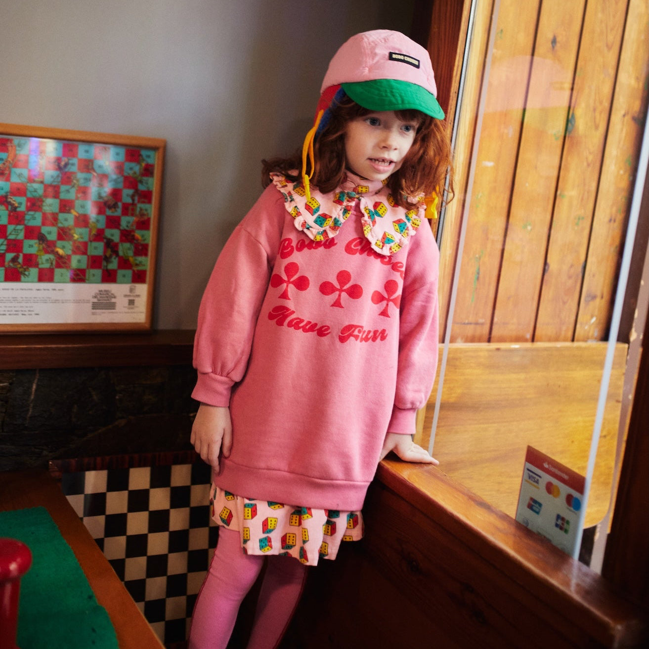 Child wearing a pink outfit with a colorful collar and hat in a room with wooden walls and a checkered floor.