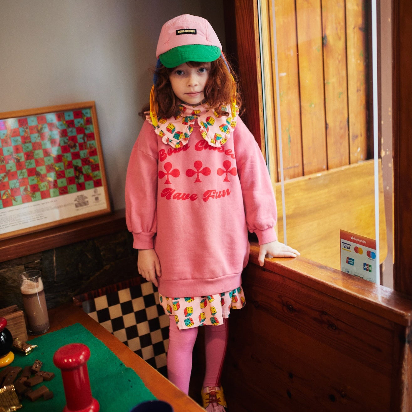 Young girl in a pink outfit standing in a room with wooden walls and a checkered floor.