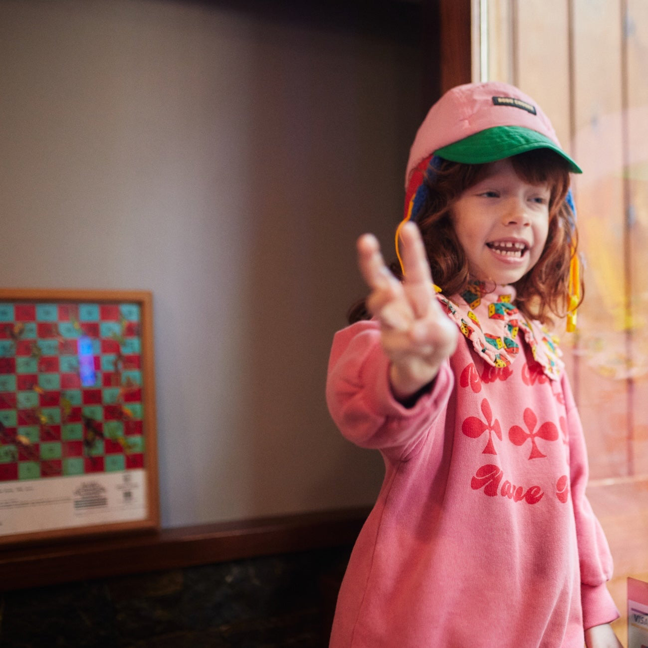 Child in pink outfit and cap making a peace sign indoors
