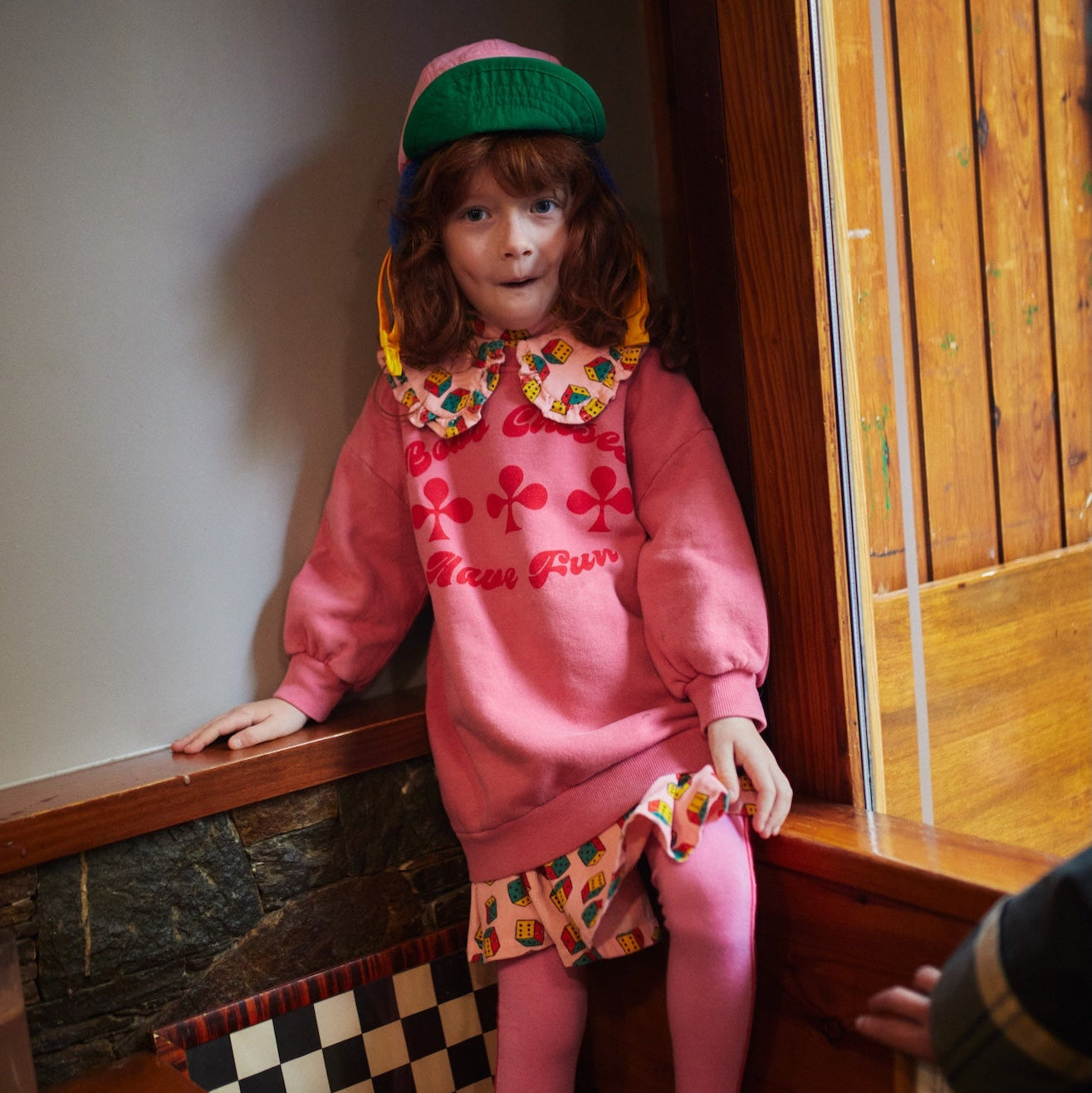 Person in a pink outfit with a colorful collar sitting on a wooden ledge.