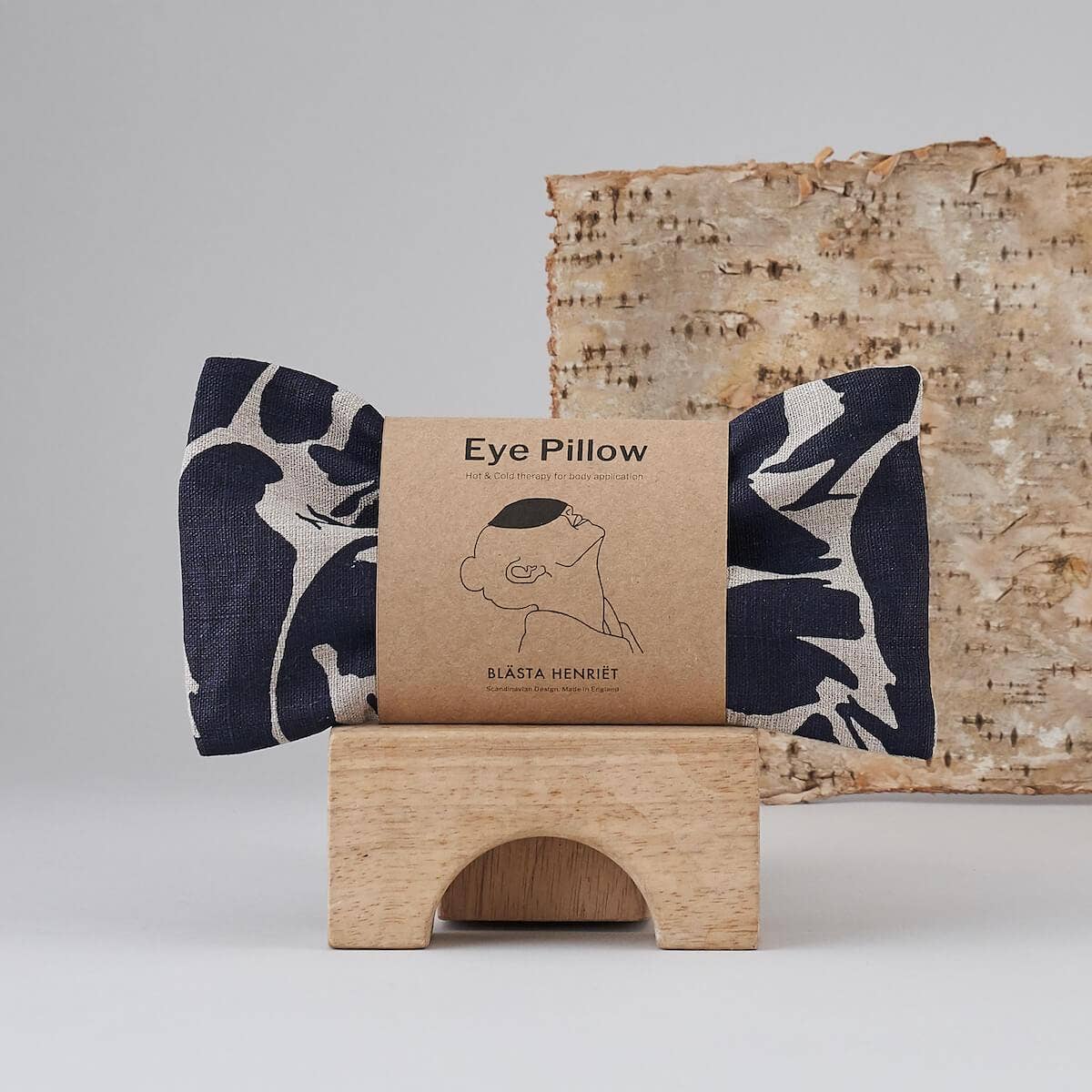 Blästa Henriët eye pillow with a navy and white abstract print, displayed in eco-friendly packaging on a wooden stand with a natural birch backdrop
