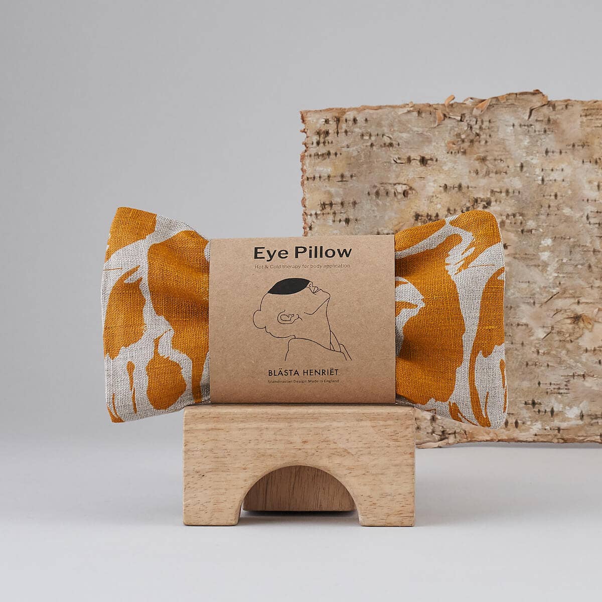 Blästa Henriët eye pillow with a bold yellow and white abstract print, displayed in eco-friendly packaging on a wooden stand with a natural birch backdrop.