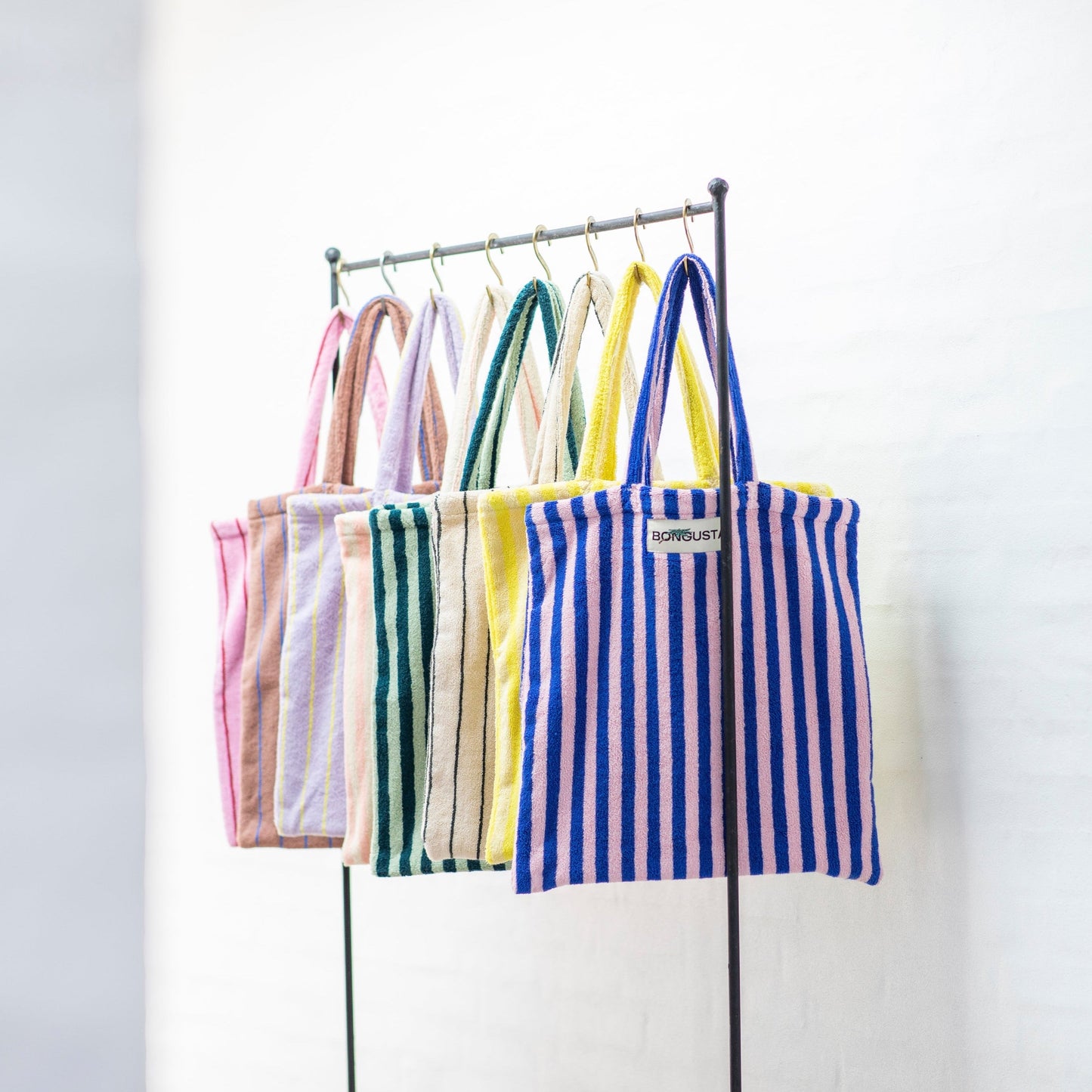 8 Bongusta classic tote bags hang on rustic gold hangers on a black rail. The bags are pink and thin neon striped, brown and thin blue striped, lilac and thin neon yellow striped, cream and pink striped, light and dark green striped, cream and thin black striped, cream and neon yellow striped and a pink and neon blue striped bag.