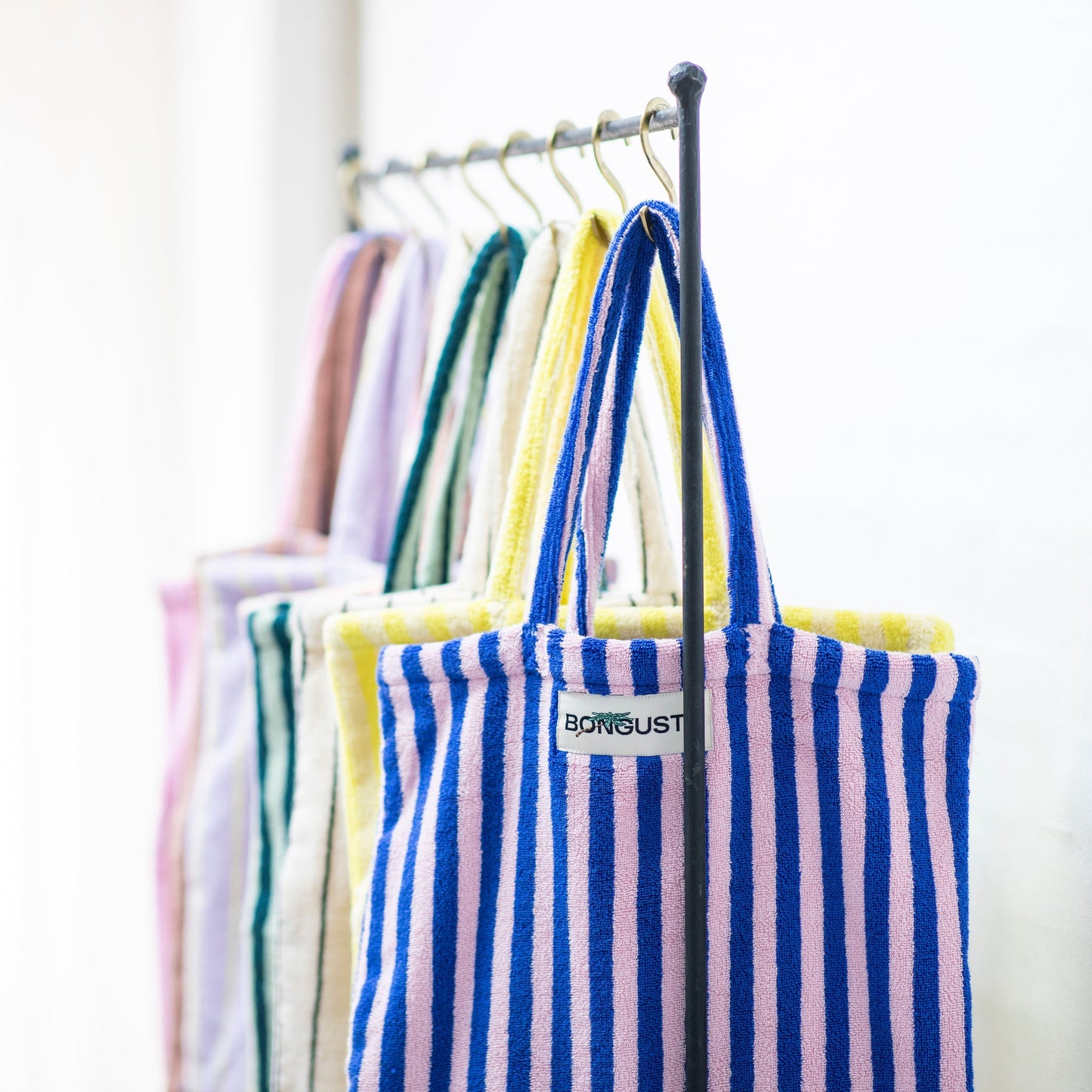 8 Bongusta classic tote bags hang on rustic gold hangers on a black rail. The bags are pink and thin neon striped, brown and thin blue striped, lilac and thin neon yellow striped, cream and pink striped, light and dark green striped, cream and thin black striped, cream and neon yellow striped and a pink and neon blue striped bag.