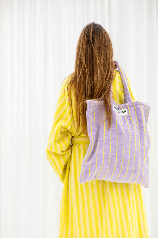 A lilac and thin neon yellow striped classic tote bag, made of towelling fabric with a lilac and thin neon yellow striped handle is held in one hand over someones back who is wearing a cream and neon yellow striped towelling dressing gown. At the top of the bag it says BONGUSTA in black inside of a cream box with green and lilac flowers coming off the words bongusta.