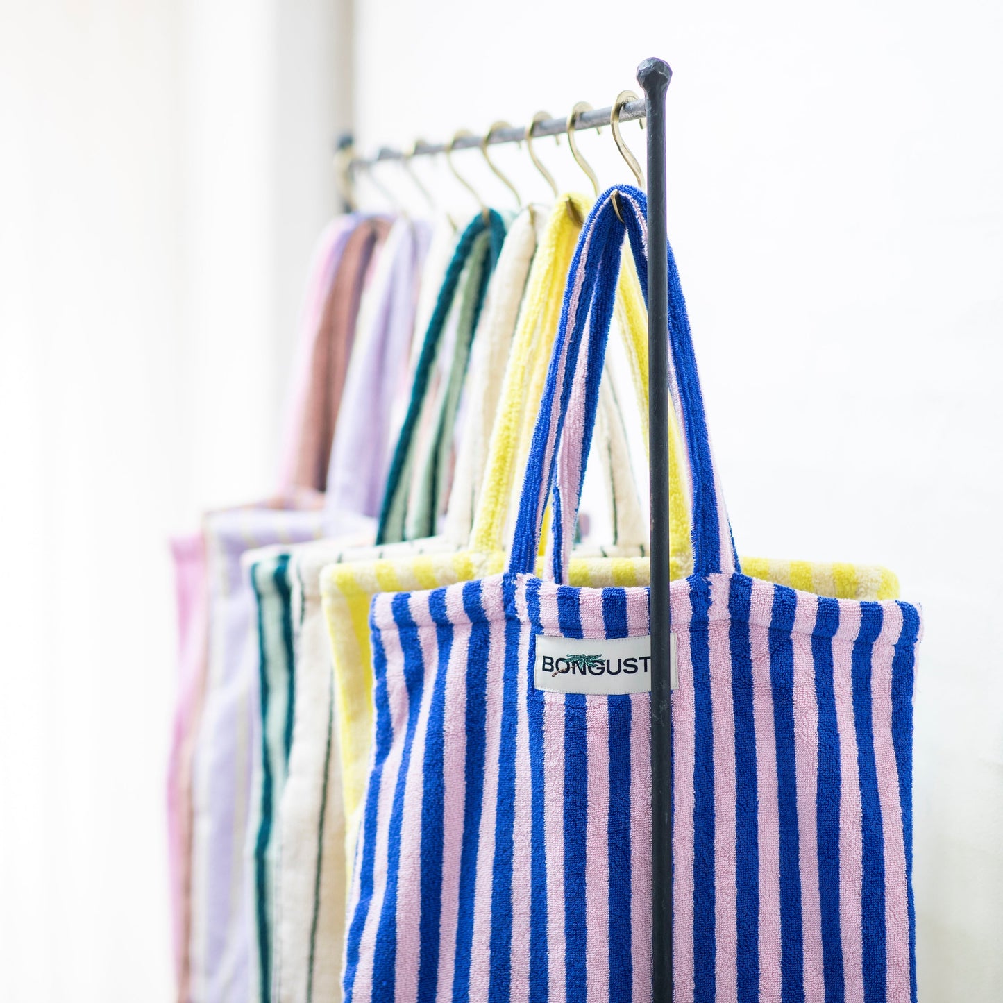8 Bongusta classic tote bags hang on rustic gold hangers on a black rail. The bags are pink and thin neon striped, brown and thin blue striped, lilac and thin neon yellow striped, cream and pink striped, light and dark green striped, cream and thin black striped, cream and neon yellow striped and a pink and neon blue striped bag.