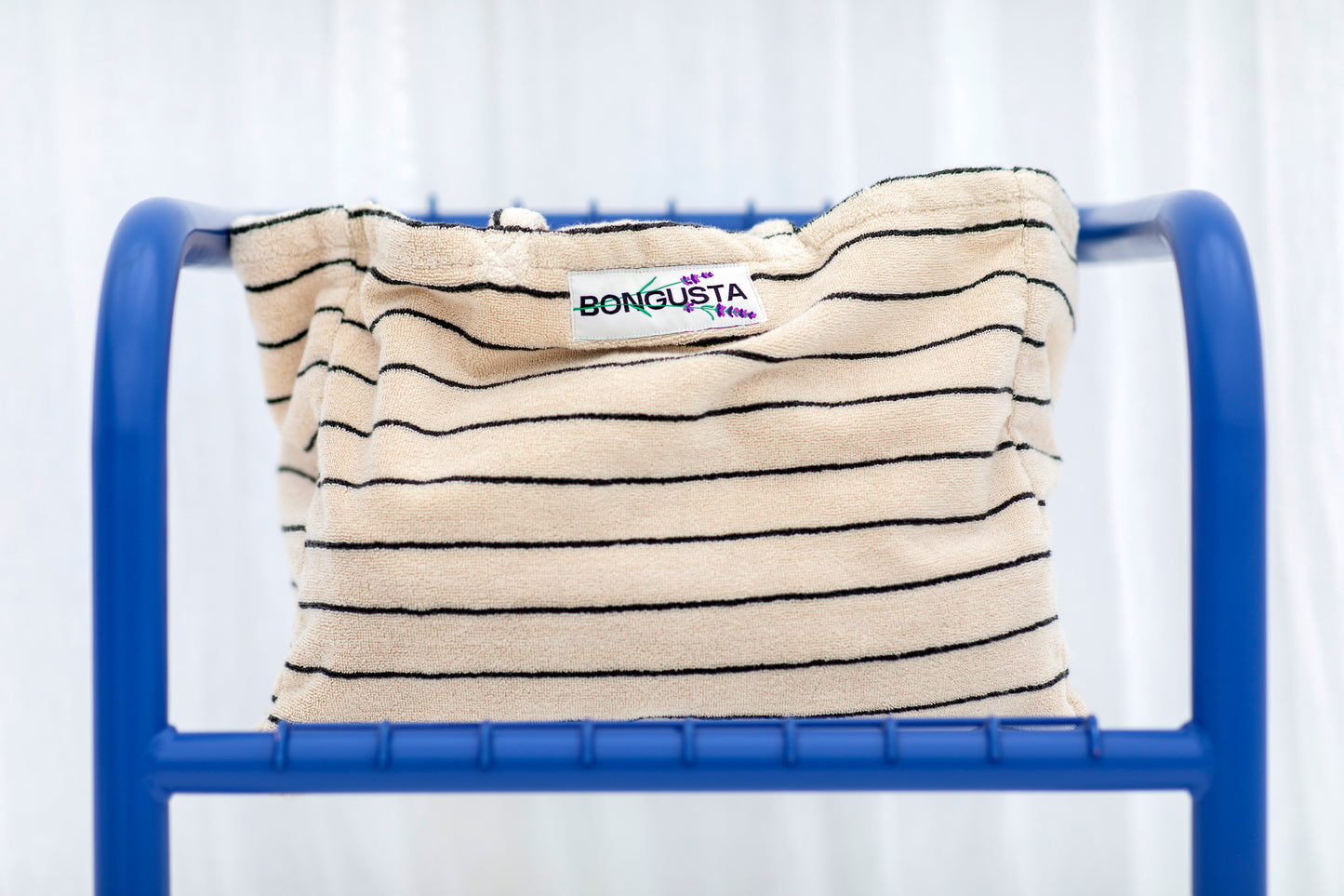 A cream weekend style tote bag with thin black stripes, made of towelling fabric with a long cream and black striped handle sits on a bright blue chair. At the top of the bag it says BONGUSTA in black with green flowers coming off the letters, inside of a cream box.