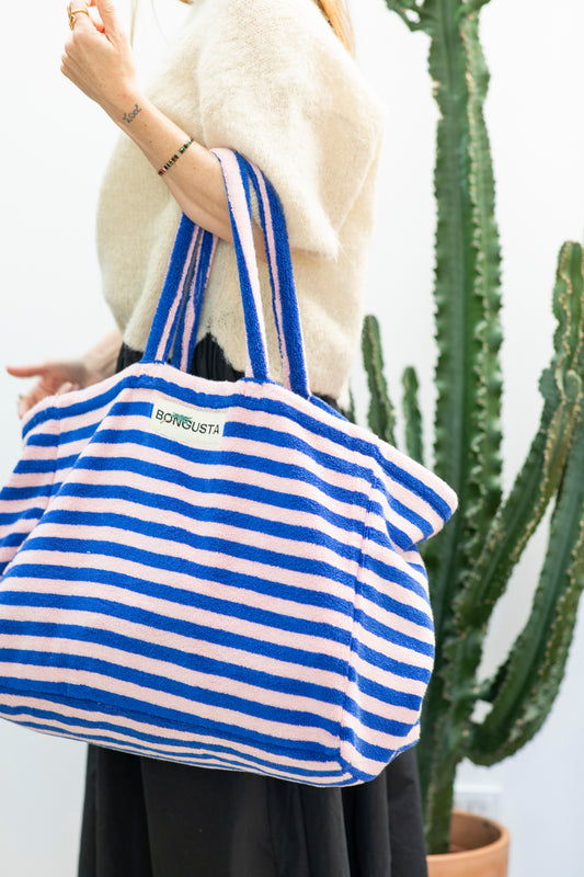 A light pink and dark blue weekend style tote bag with thin red stripes, made of towelling fabric with a long pink and blue striped handle is held over there arm by someone wearing a cream jumper and black wide legged trousers in front of a cactus. At the top of the bag it says BONGUSTA in black with green flowers coming off the letters, inside of a cream box.