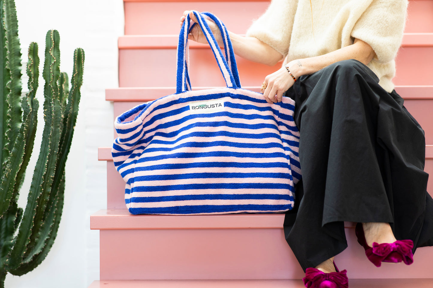 A light pink and dark blue weekend style tote bag with thin red stripes, made of towelling fabric with a long pink and blue striped handle sits on baby pink steps next to someone wearing a cream jumper and wide legged black trousers with pink and velvet flats on that have bows on top and on the other side stands a green cactus. At the top of the bag it says BONGUSTA in black with green flowers coming off the letters, inside of a cream boxes.
