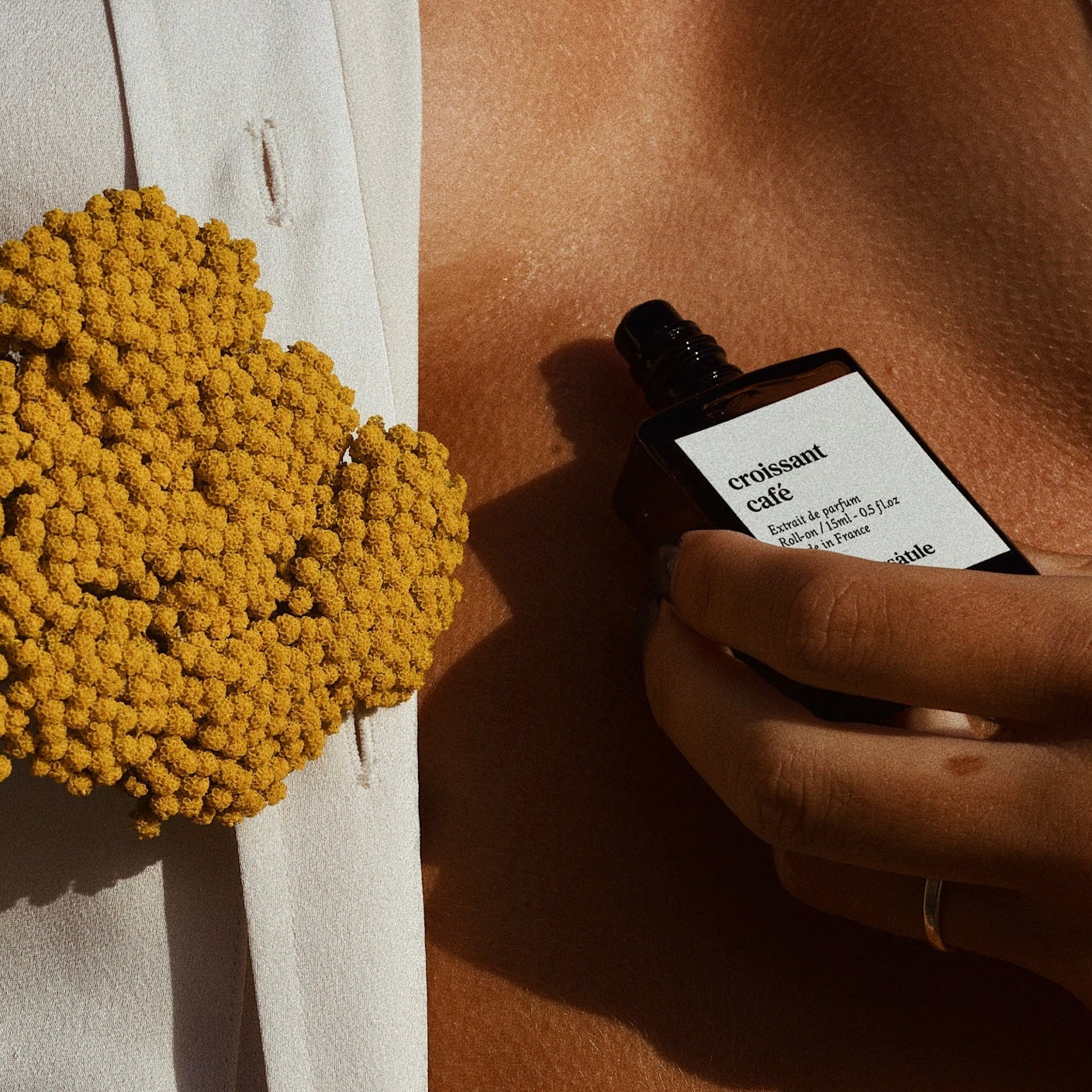 Hand holding a perfume bottle labeled 'cruissant cafe' next to a yellow floral arrangement.