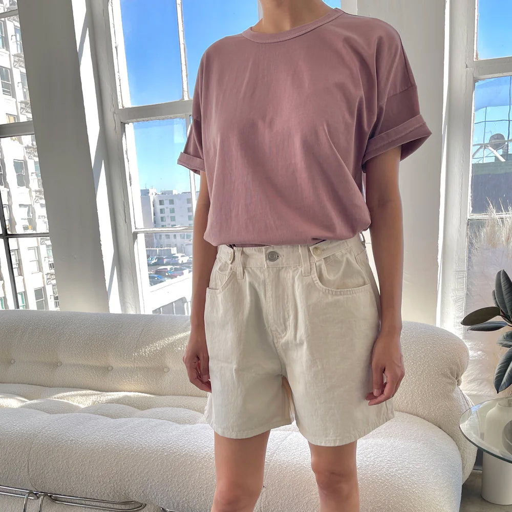 A person wearing a pink crew neck t-shirt with rolled-up sleeves and a pair of beige shorts, standing in a room with large windows.