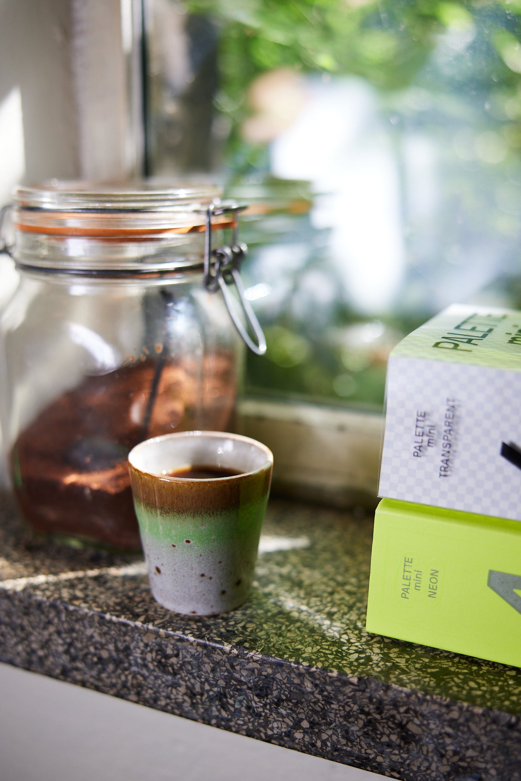 A hkliving good vibes ristretto mug (1 of 4) with a white speckled body and a faded lime green stripe which fades into a warm brown trim, filled with black coffee. All on a grey marble windowsill it mug is in front of a mason jar half filled with ground coffee, and infront of the mug is two books stacked on top of eachother