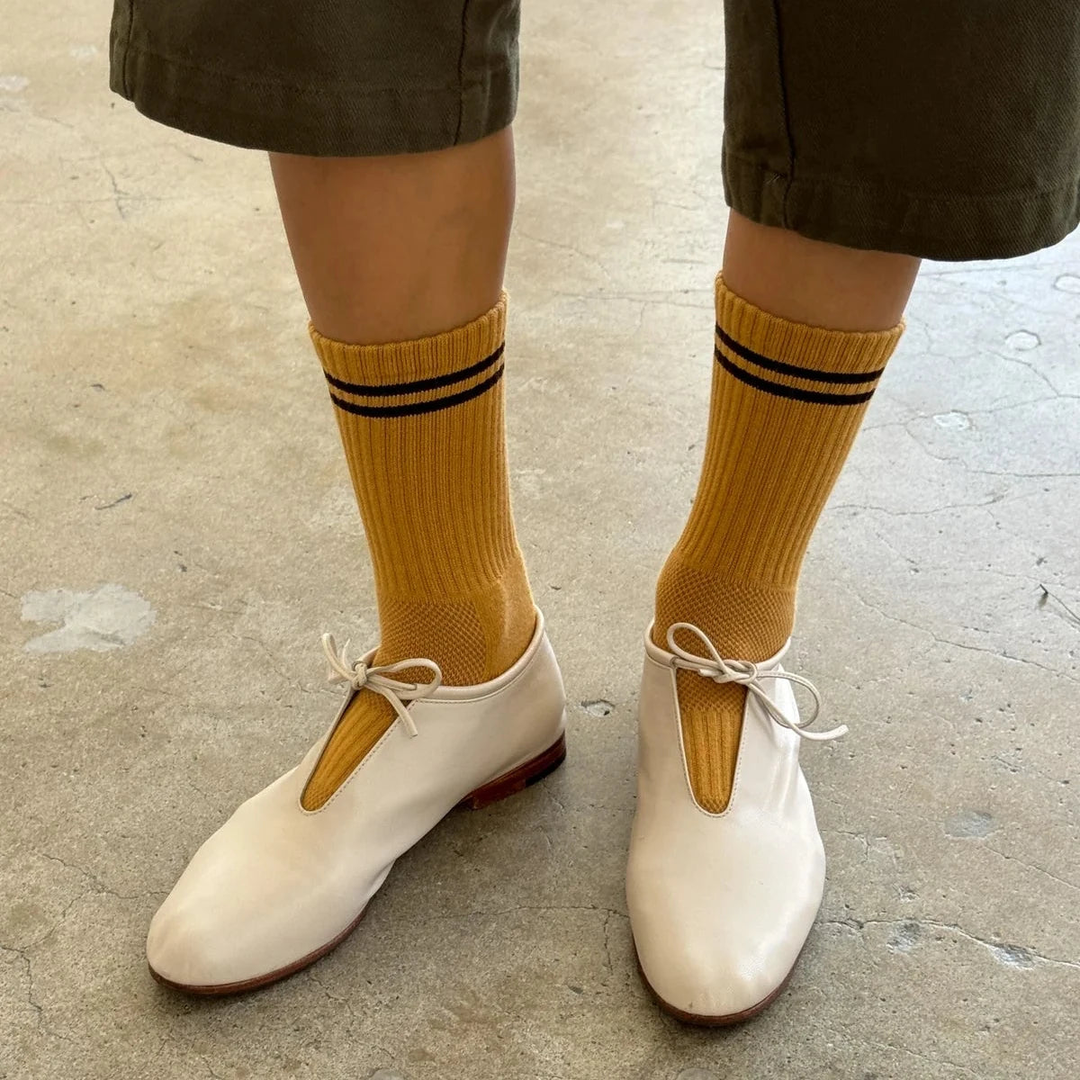 Person wearing mustard yellow socks with black stripes and white shoes on a concrete floor.