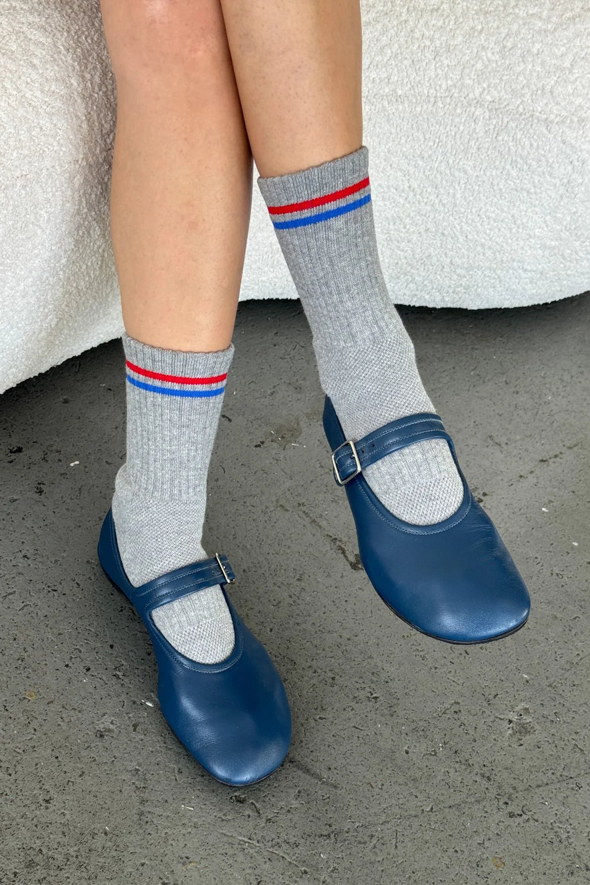Person wearing gray socks with red and blue stripes and blue shoes on a concrete floor.