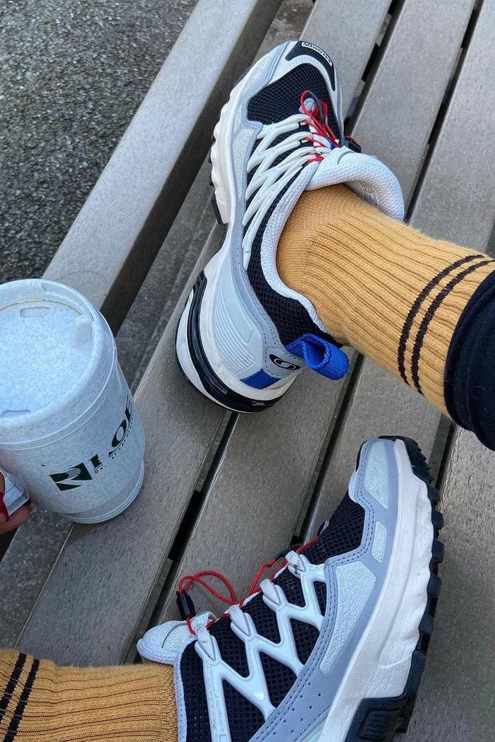 Person wearing white sneakers with black and blue accents on a wooden bench, next to a white cup.